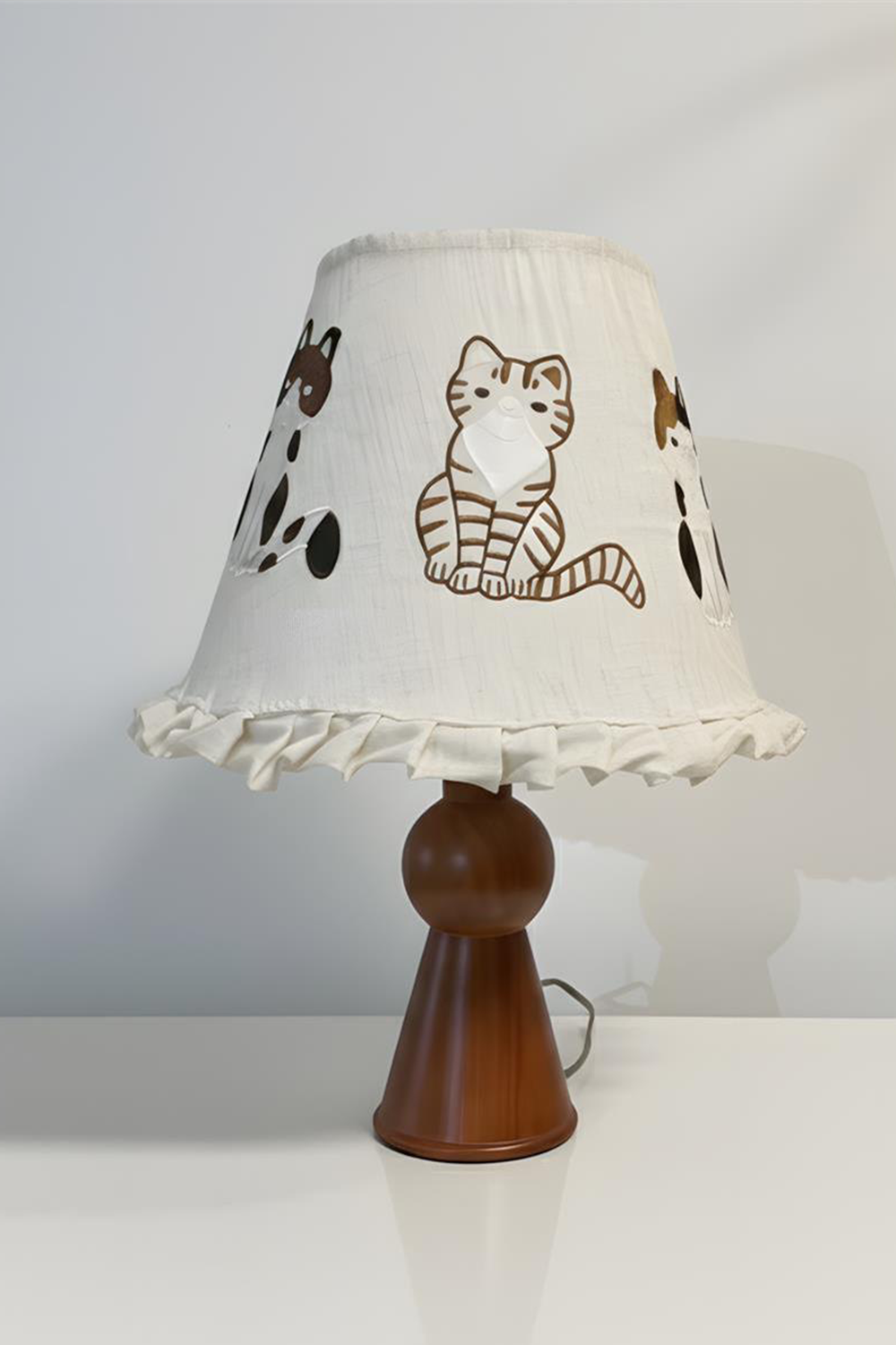 Whiskers Ruffled Fabric Table Lamp | Fabric, Wood | Front view of the fully lit embroidered cat ruffle lamp sitting on a white table against a neutral grey studio background | Samu Lamp