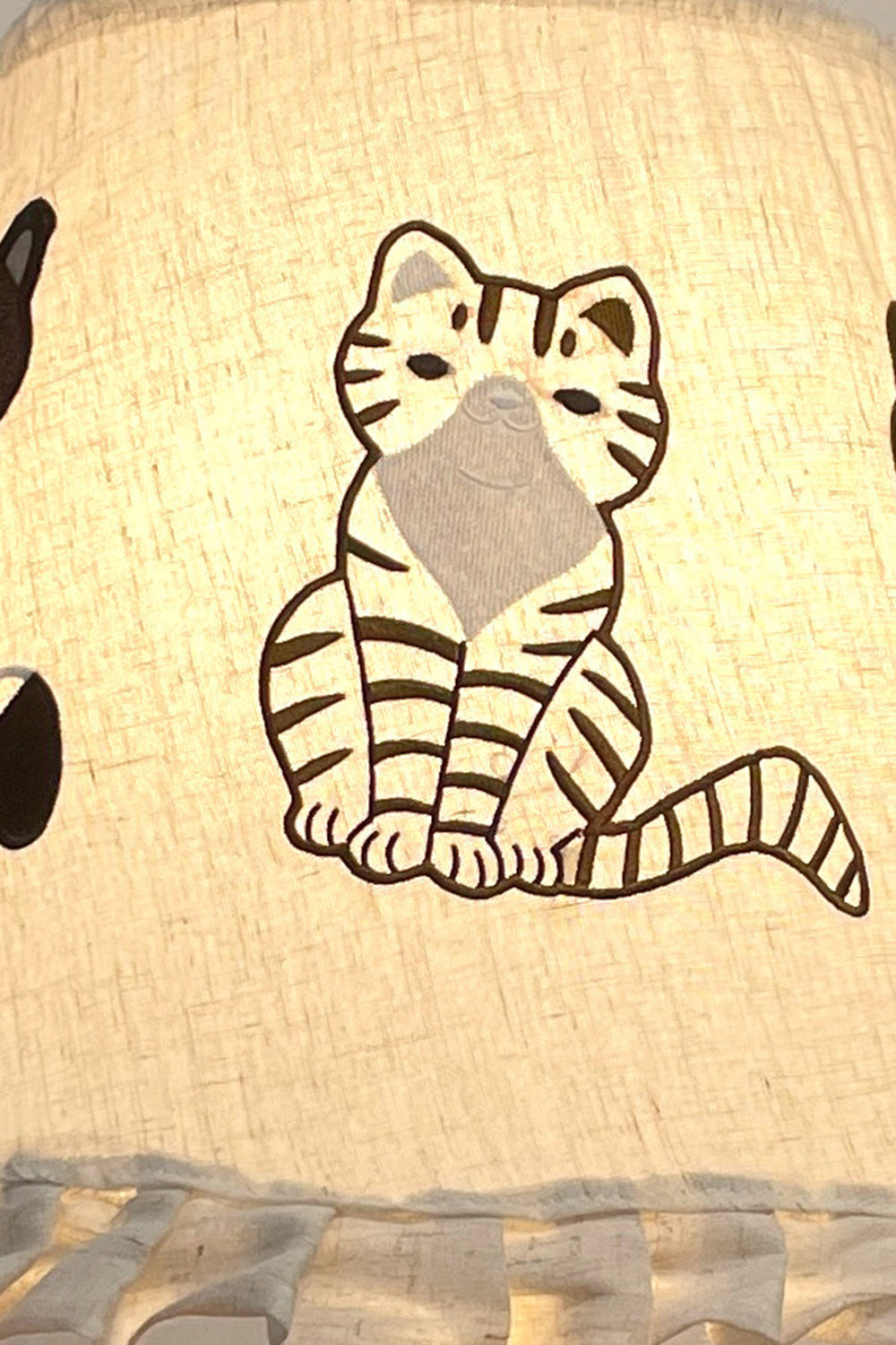 Whiskers Ruffled Fabric Table Lamp | Fabric, Wood | Macro close-up focus on the illuminated white linen shade highlighting the detailed embroidered cartoon cat pattern | Detail shot | Samu Lamp