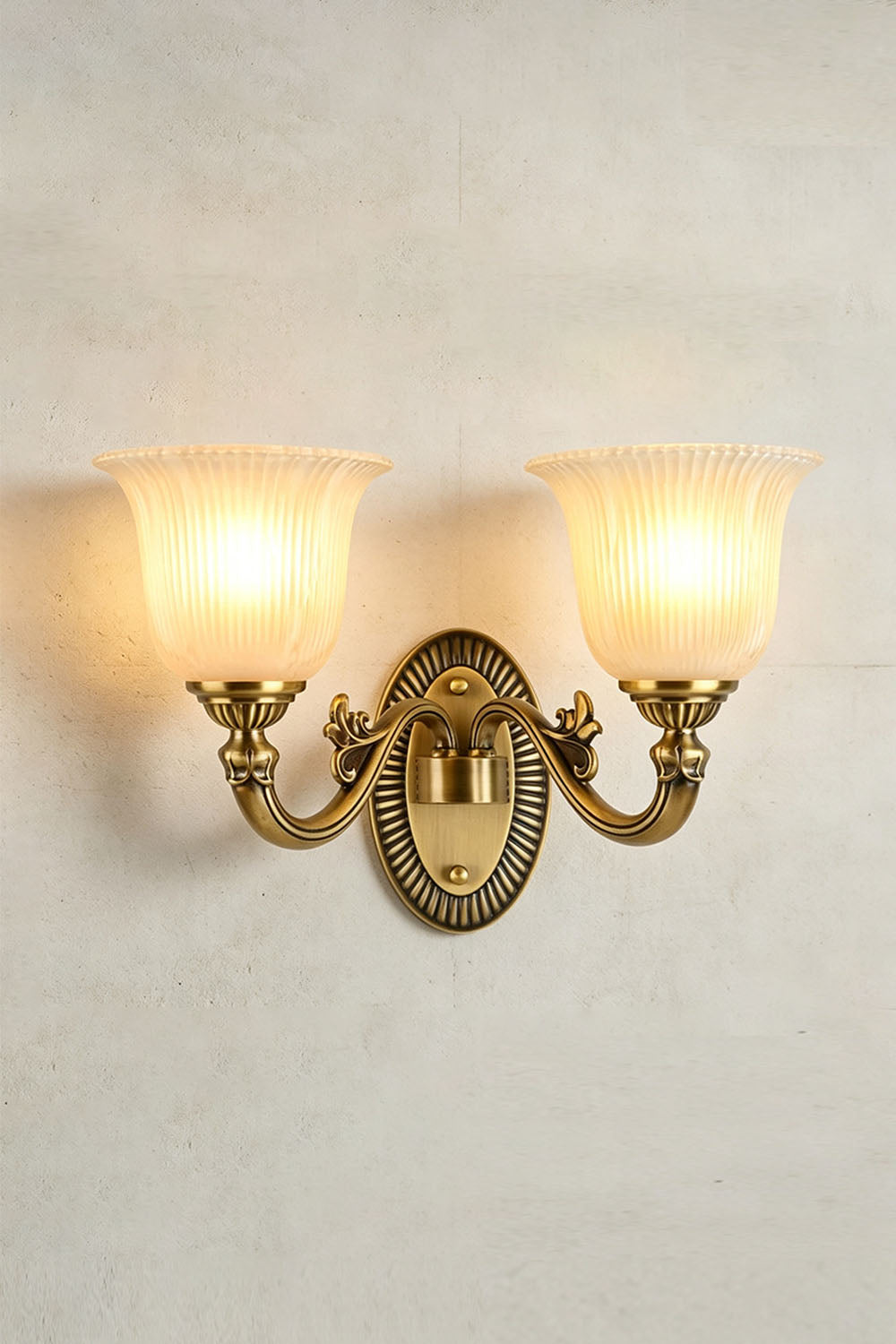 Whitlock Fluted Bell Brass Wall Lamp with two glowing shades on a textured wall.