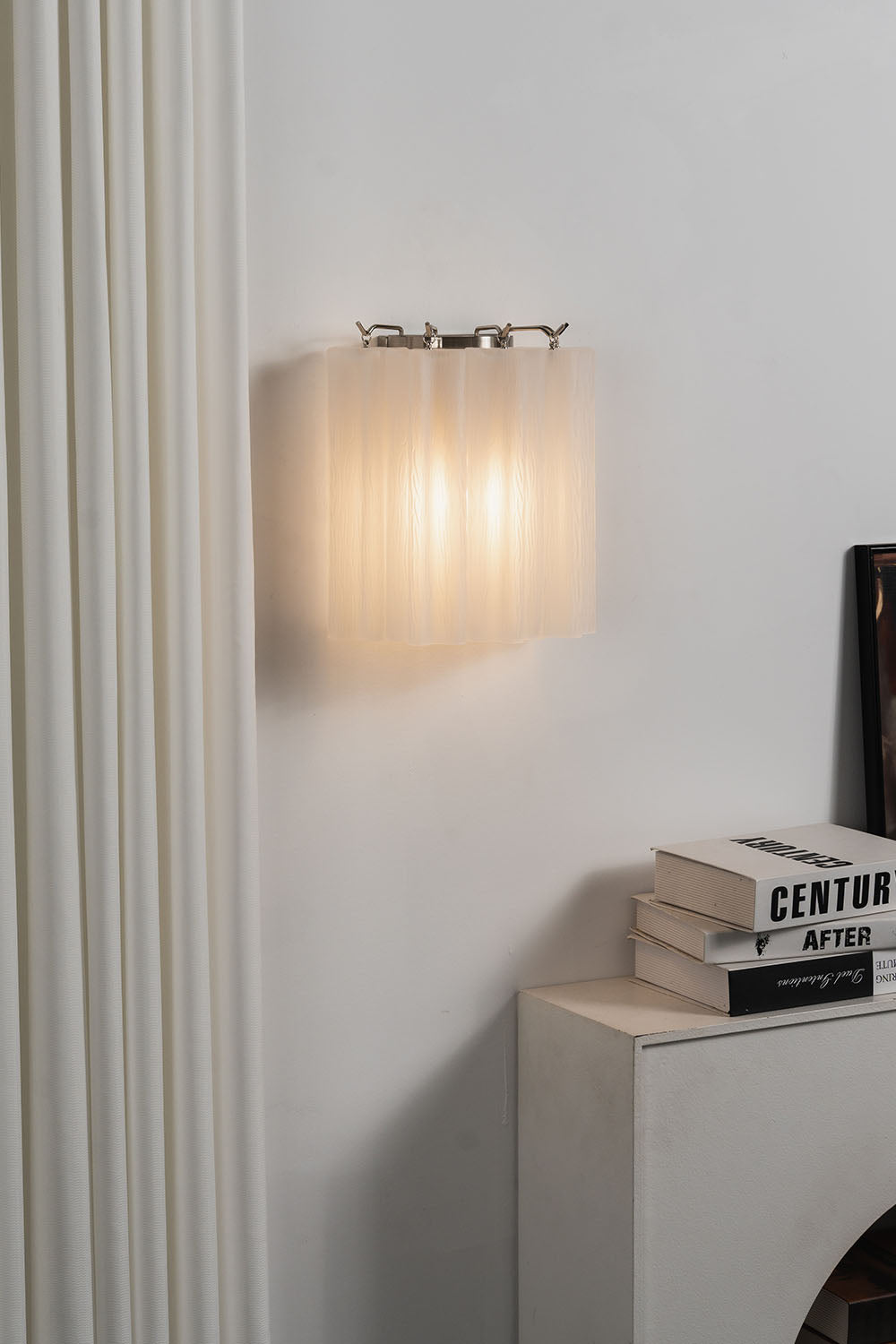 Ysoline Rippled Glass Curtain Sconce Light | Rippled Glass and Metal | Glowing softly on a wall near a small white side table with books | Samu Lamp